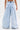 A person wearing the IN CHARGE WIDE LEG DENIM PANT, featuring a high-rise waist, light-wash, wide leg fit with large back pockets and frayed hems, stands with their back to the camera against a plain white background.