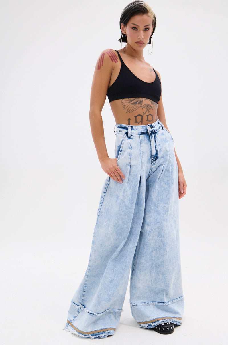 A person stands confidently in the IN CHARGE WIDE LEG DENIM PANT with distressed hemlines and a high-rise waist, paired with a black crop top. Tattoos, hoop earrings, and slicked-back wet-look hair complete their bold look.
