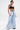 A person stands confidently in the IN CHARGE WIDE LEG DENIM PANT with distressed hemlines and a high-rise waist, paired with a black crop top. Tattoos, hoop earrings, and slicked-back wet-look hair complete their bold look.