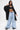 A woman exudes confidence in the IN CHARGE WIDE LEG DENIM PANT, paired with a black hooded jacket and crop top. She showcases tattoos on her torso, large earrings, and holds her hood against a plain white background.
