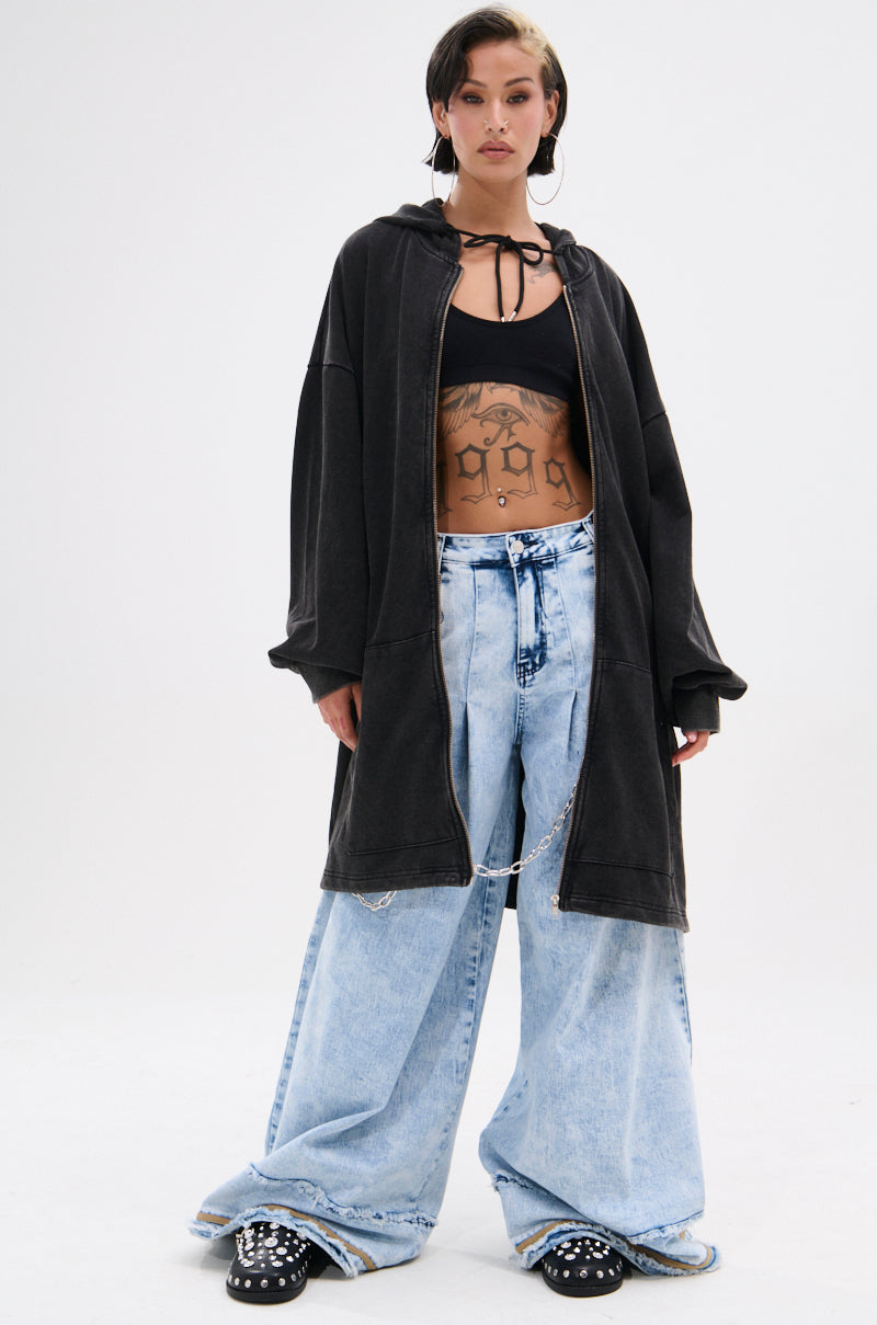 A person stands confidently in light blue IN CHARGE WIDE LEG DENIM PANT with distressed hems and a chain belt, black crop top, oversized black zip-up hoodie, and black shoes. Their midriff tattoos are visible, and they have short dark hair.