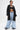 A person stands confidently in light blue IN CHARGE WIDE LEG DENIM PANT with distressed hems and a chain belt, black crop top, oversized black zip-up hoodie, and black shoes. Their midriff tattoos are visible, and they have short dark hair.