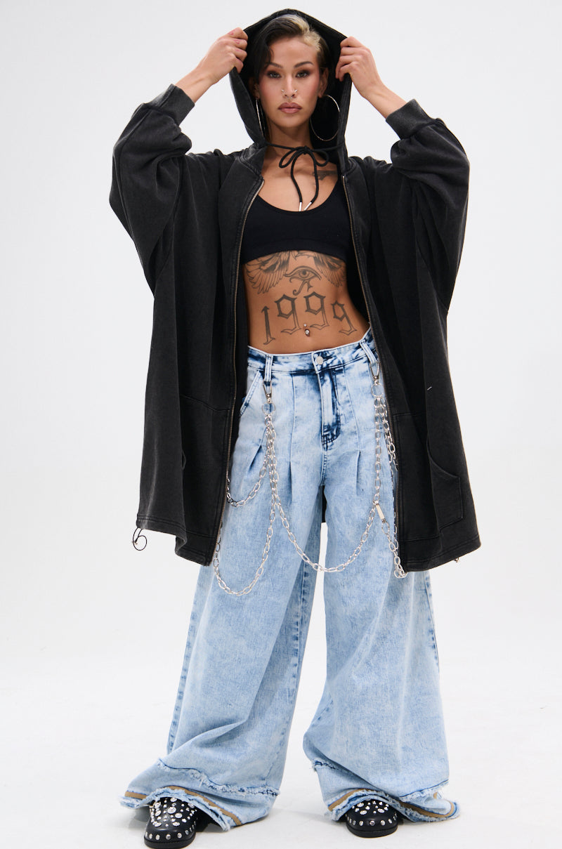 A woman poses in the IN CHARGE WIDE LEG DENIM PANT, paired with a black hooded oversized jacket and crop top. She holds the hood with both hands, showing tattoos on her torso. The jeans feature light blue denim, distressed hems, and silver chains.