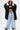 A woman poses in the IN CHARGE WIDE LEG DENIM PANT, paired with a black hooded oversized jacket and crop top. She holds the hood with both hands, showing tattoos on her torso. The jeans feature light blue denim, distressed hems, and silver chains.