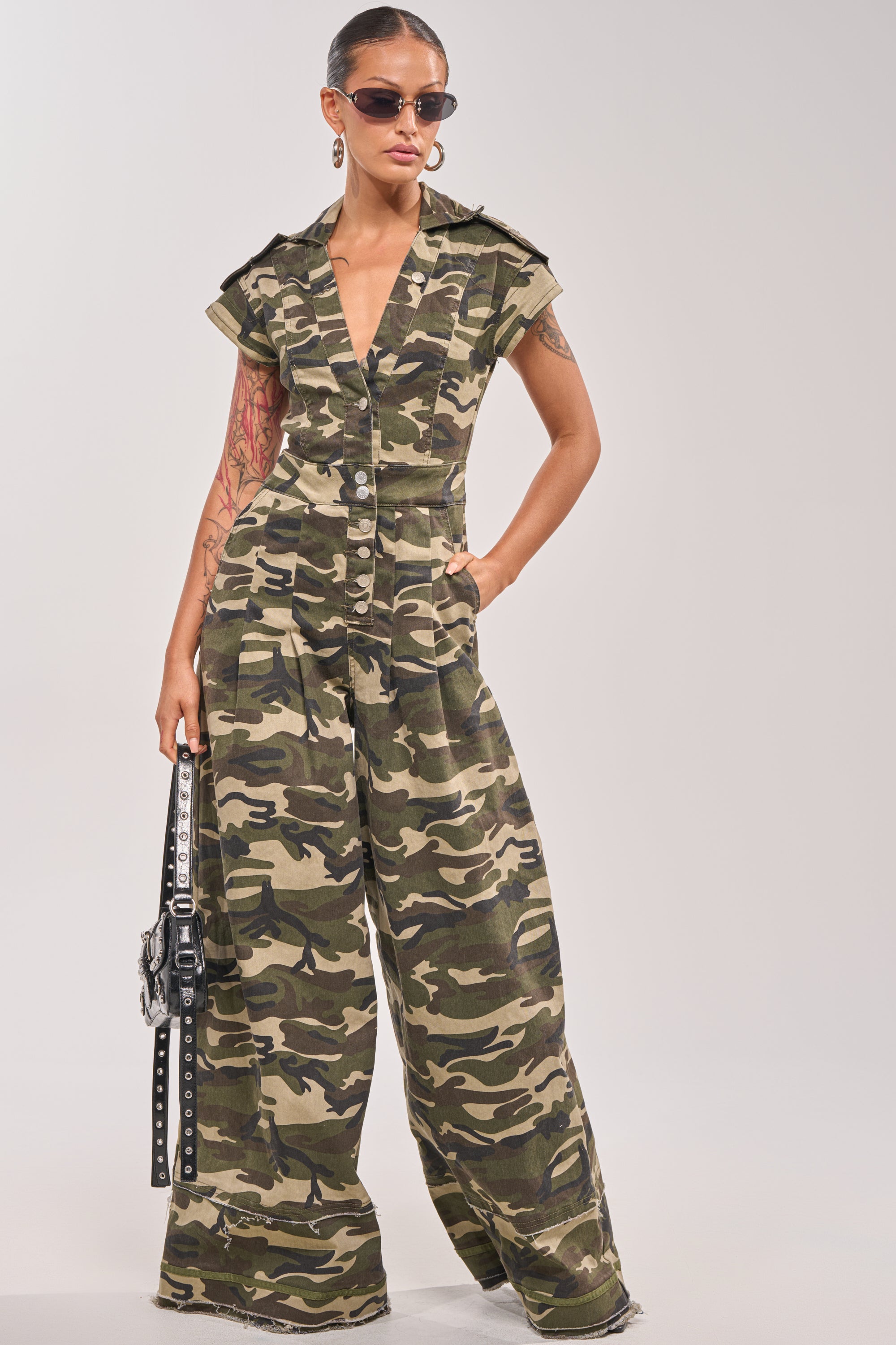 A woman confidently poses in the MAJOR DRAMA CAMO JUMPSUIT IN CAMO, styled with sunglasses, earrings, and a black handbag, her hand casually in her pocket. 0