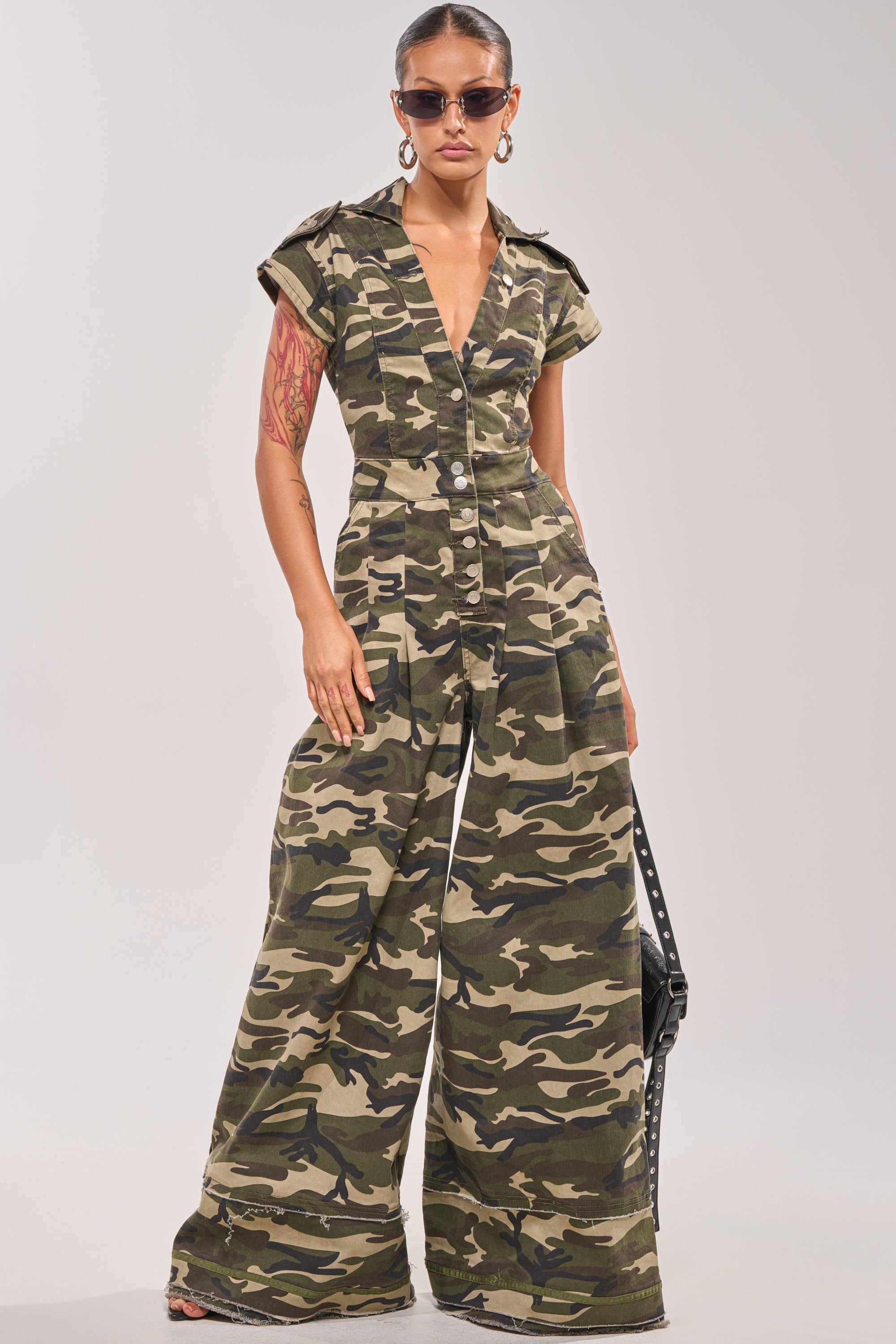 Wearing the MAJOR DRAMA CAMO JUMPSUIT IN CAMO, sunglasses, hoop earrings, and holding a studded black bag, a person stands confidently against a plain background. 1