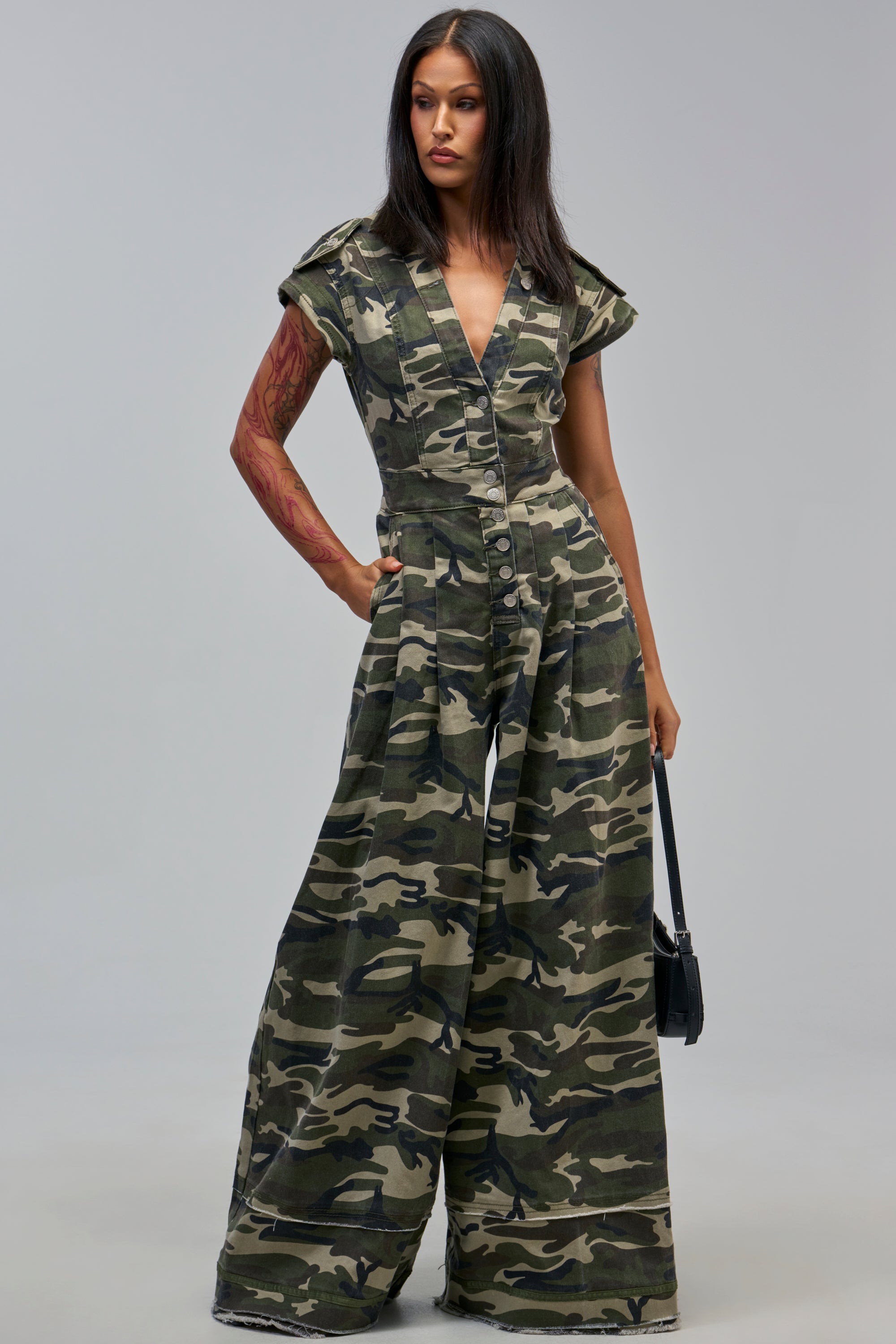 A woman in the MAJOR DRAMA CAMO JUMPSUIT IN CAMO with wide legs and cap sleeves stands holding a black bag, gazing slightly to the side.
