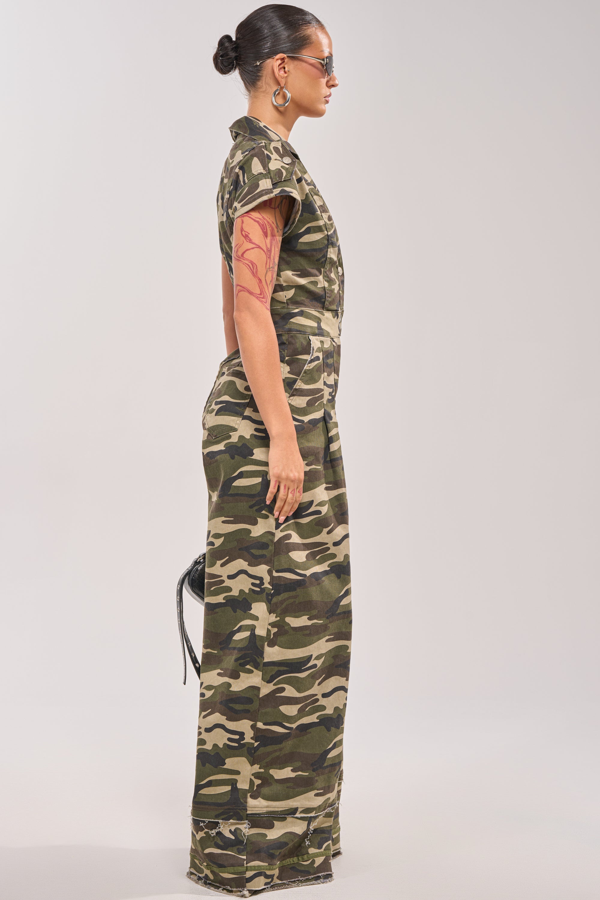 A woman in profile wears the MAJOR DRAMA CAMO JUMPSUIT IN CAMO with sunglasses, hoop earrings, an upper arm tattoo, hair in a bun, and holds a small black purse against a light gray background. 2
