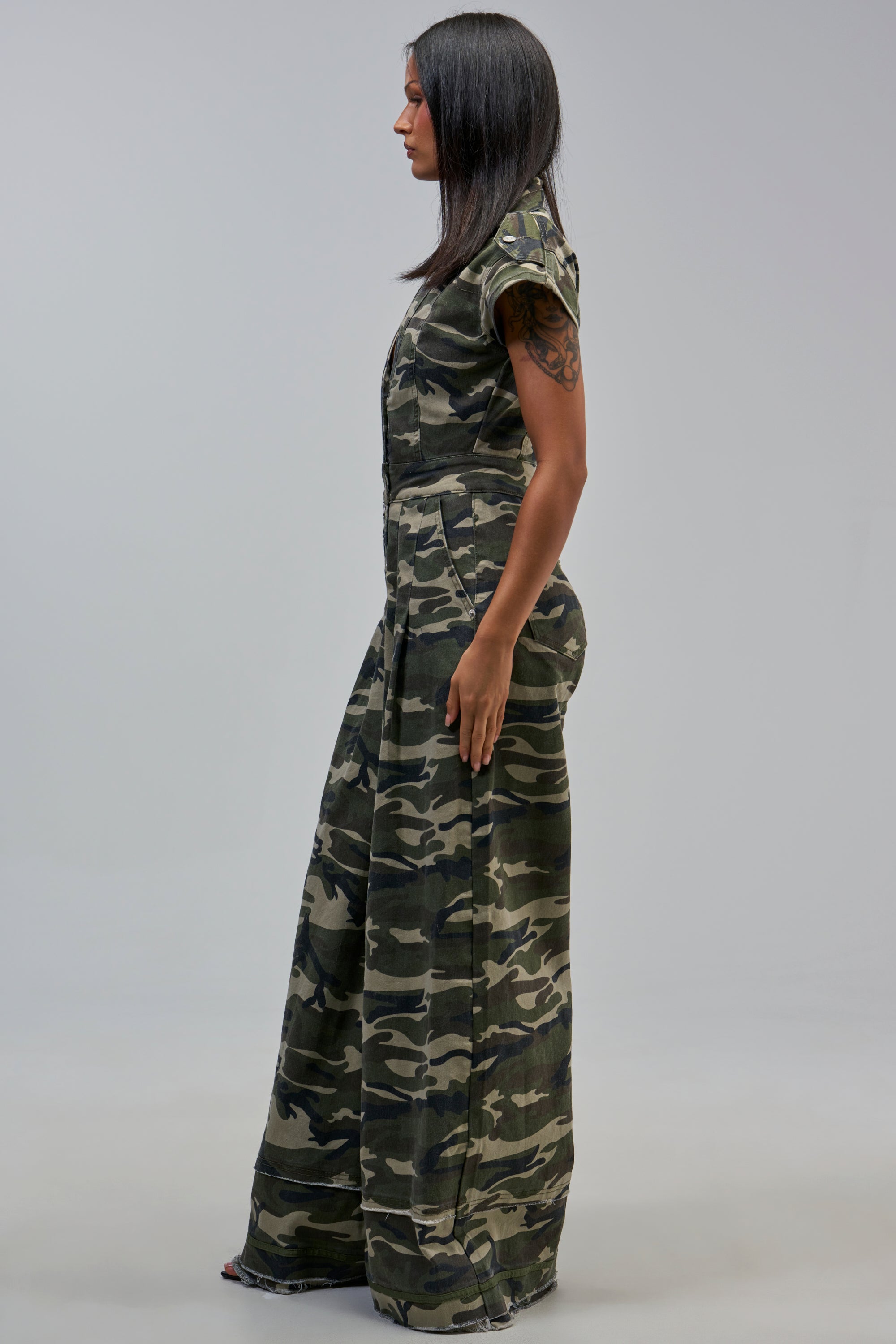 Woman in profile against a plain background wears the MAJOR DRAMA CAMO JUMPSUIT IN CAMO with black shoes, straight dark hair, and a visible upper arm tattoo.
