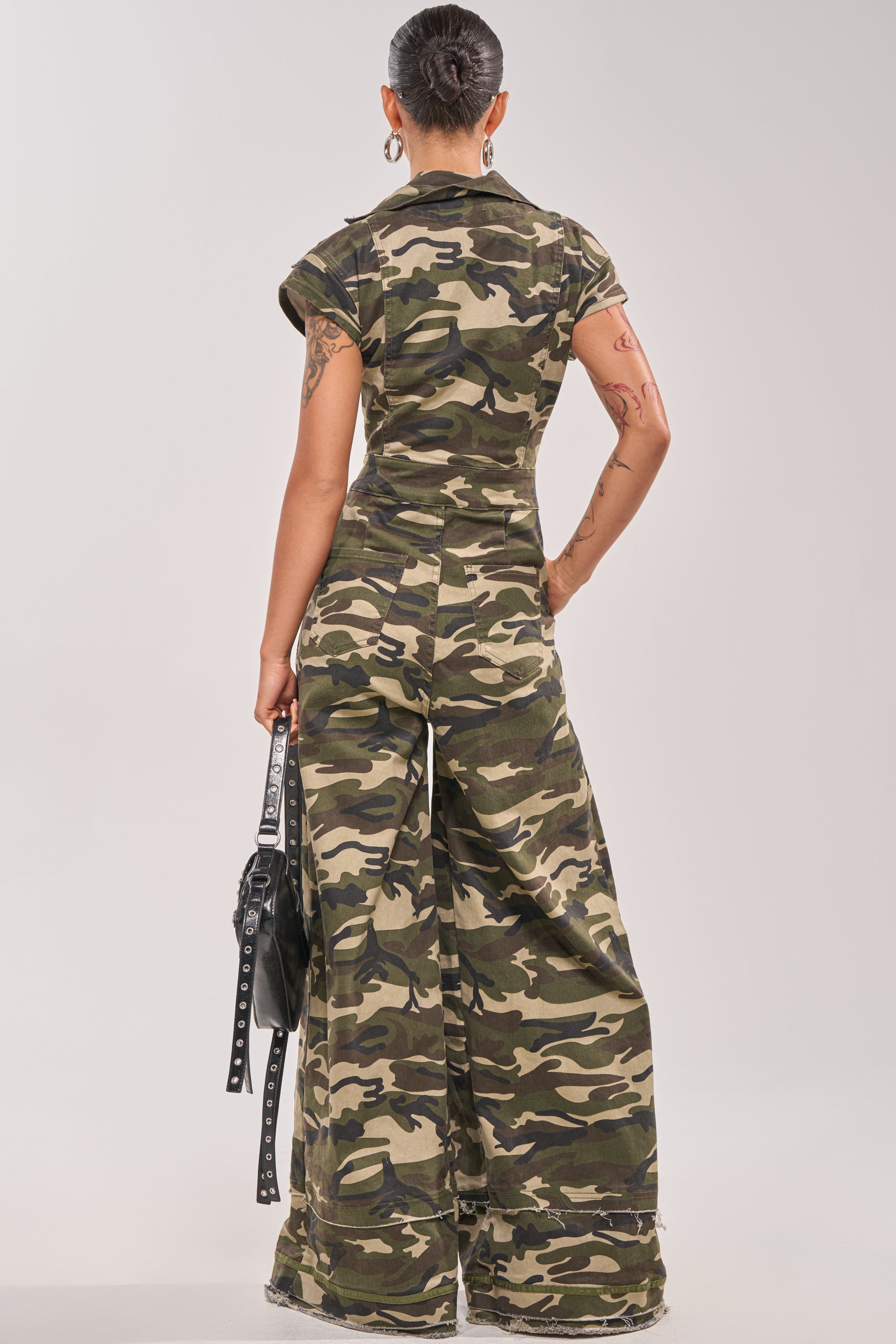 A person with short hair faces away, wearing the MAJOR DRAMA CAMO JUMPSUIT IN CAMO with wide legs and cap sleeves, holding a black handbag. Tattoos are visible on both arms against a plain, light-colored background. 3