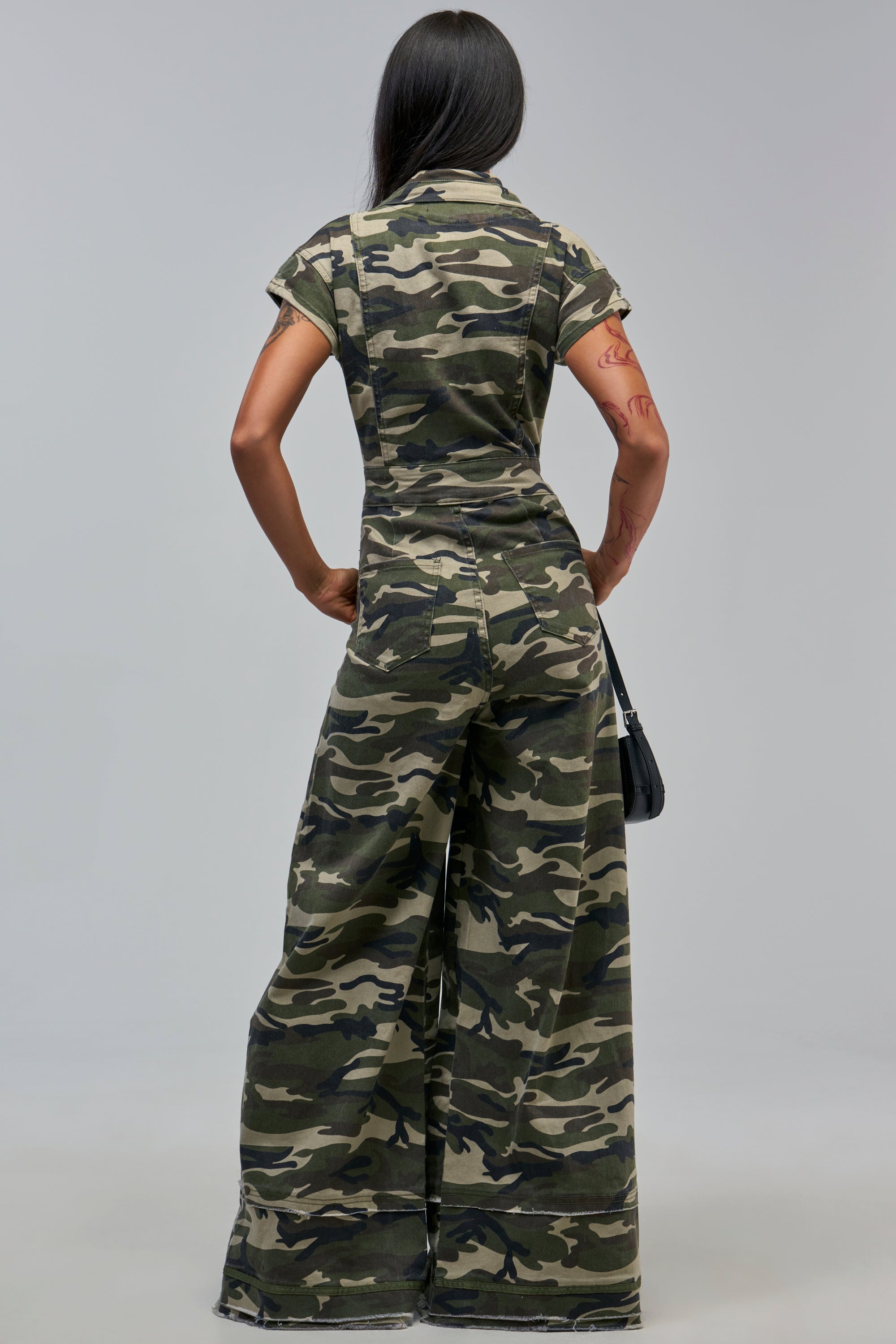 Person facing away in the MAJOR DRAMA CAMO JUMPSUIT IN CAMO—short sleeves, wide legs. They have long dark hair, a visible arm tattoo, hold a small black bag. Plain light gray background.