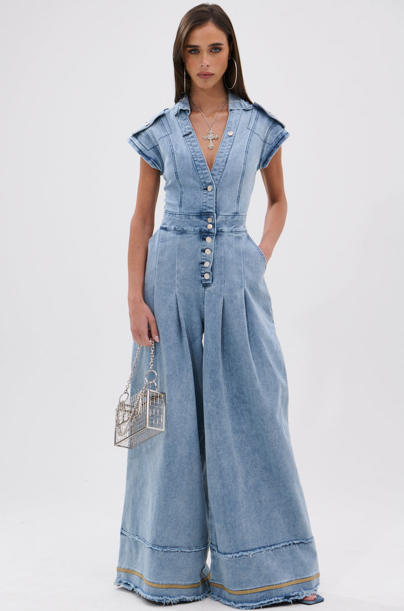 Woman models the AT LEAST I LOOK CUTE DENIM JUMPSUIT—a light-wash, wide-leg style with front buttons and a deep V-neck—styled with layered necklaces, large hoops, and a silver cage handbag against a plain white backdrop.