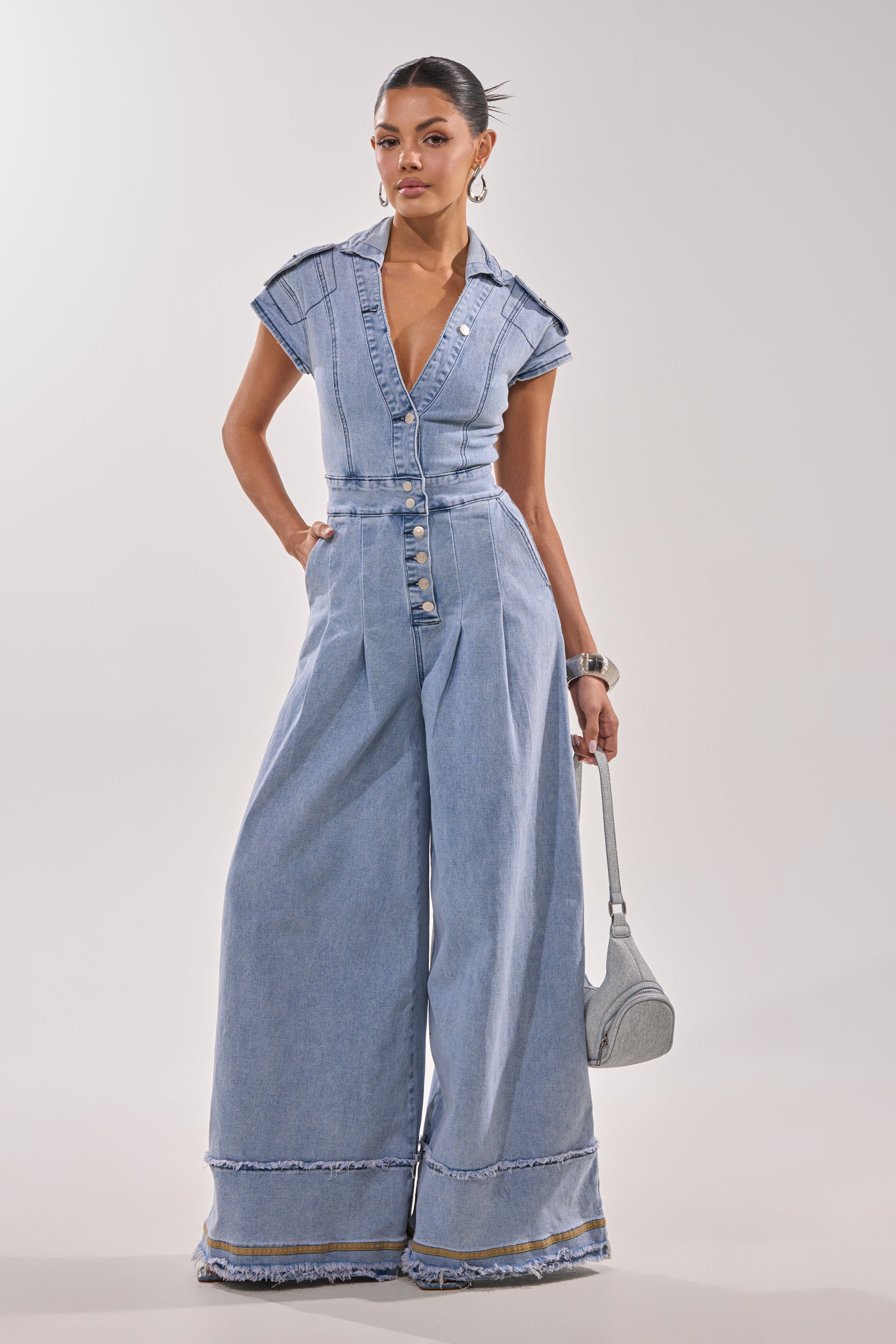 A woman stands confidently in the AT LEAST I LOOK CUTE DENIM JUMPSUIT, featuring light-wash denim, short sleeves, deep V-neck, wide-leg pants, and silver buttons. She pairs it with a small gray purse, hoop earrings, and a bangle.