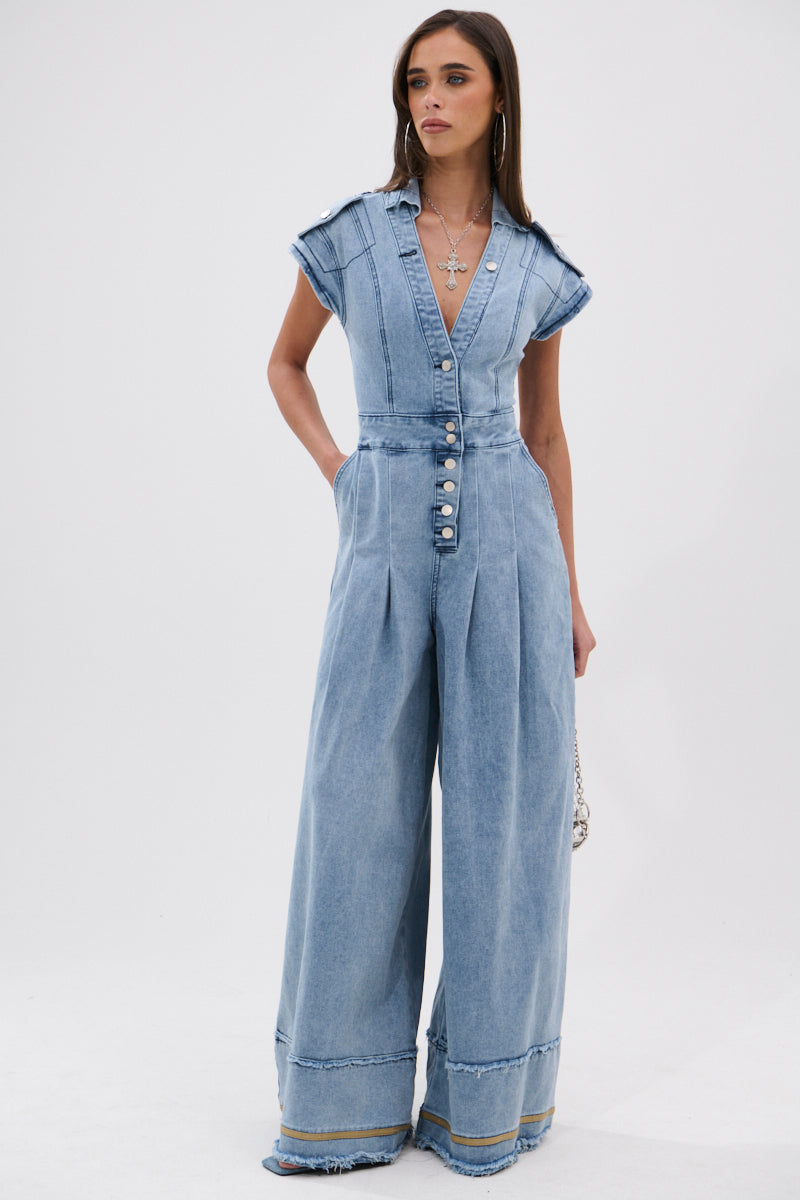A woman with long hair models the AT LEAST I LOOK CUTE DENIM JUMPSUIT, a light-wash style featuring wide legs, short sleeves, a deep V-neck, and button details. She stands against a plain white background with a small bag for an effortless look.