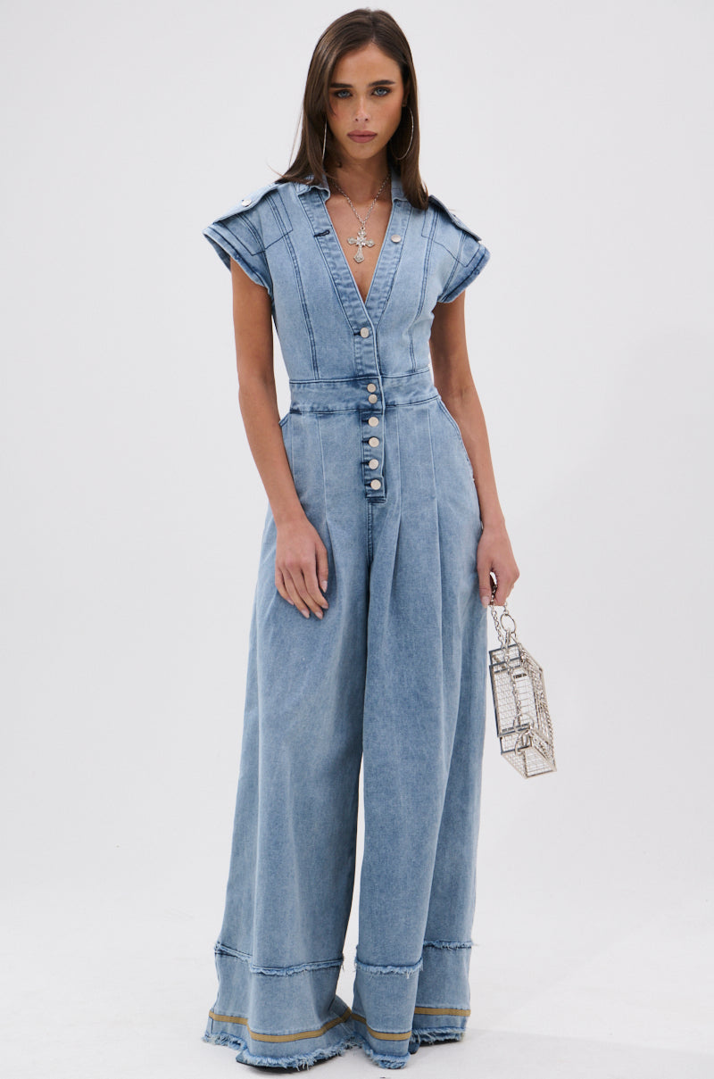 A woman poses against a white background wearing the AT LEAST I LOOK CUTE DENIM JUMPSUIT, featuring a light wash, cap sleeves, deep V-neck, and wide legs. She accessorizes with a metallic box handbag and layered necklaces for utility-chic style.