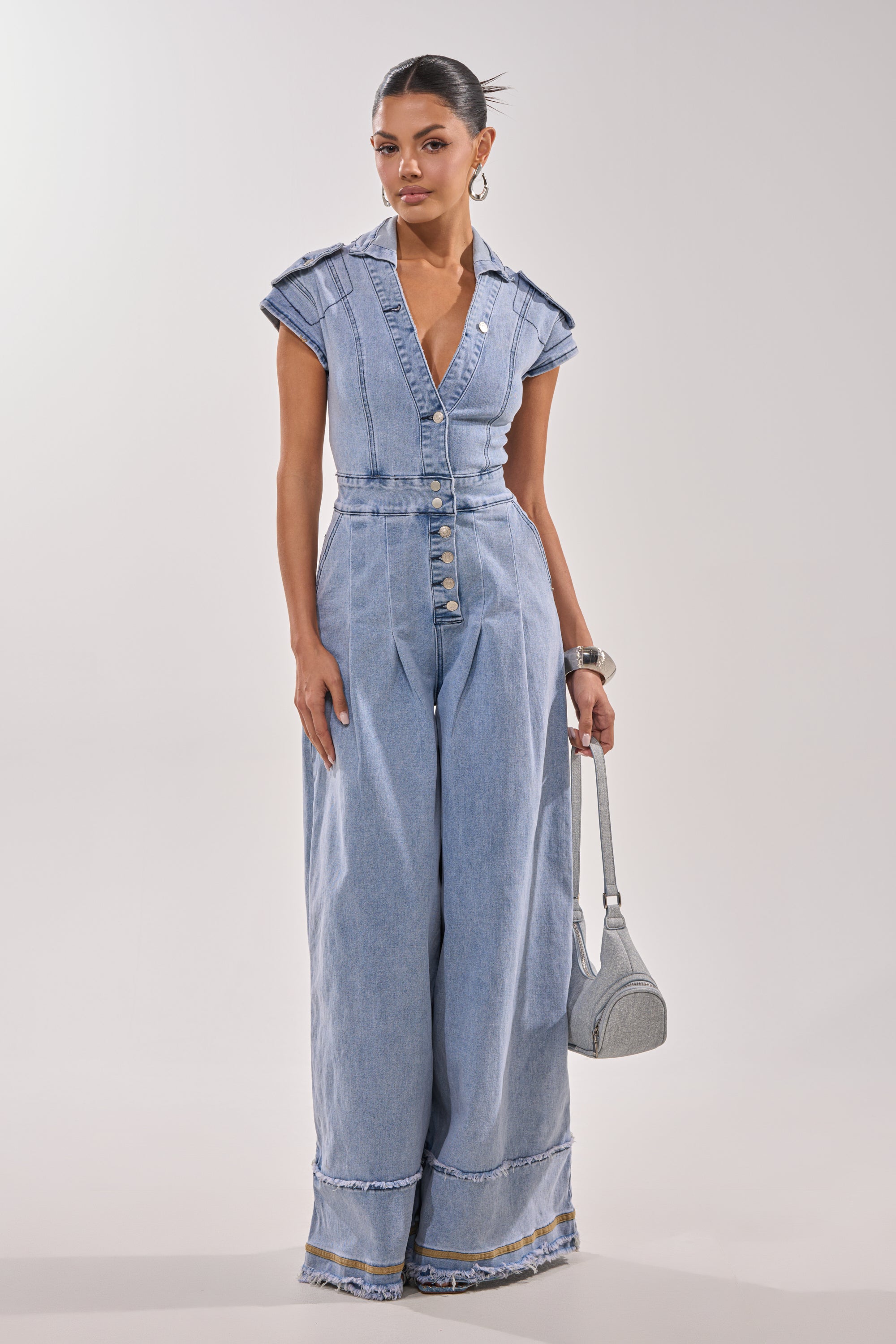 A woman poses against a plain background in the AT LEAST I LOOK CUTE DENIM JUMPSUIT, a utility-inspired, light blue, wide-leg piece with cap sleeves, a deep V-neckline, and front buttons. She accessorizes with hoop earrings and a small gray handbag. 2