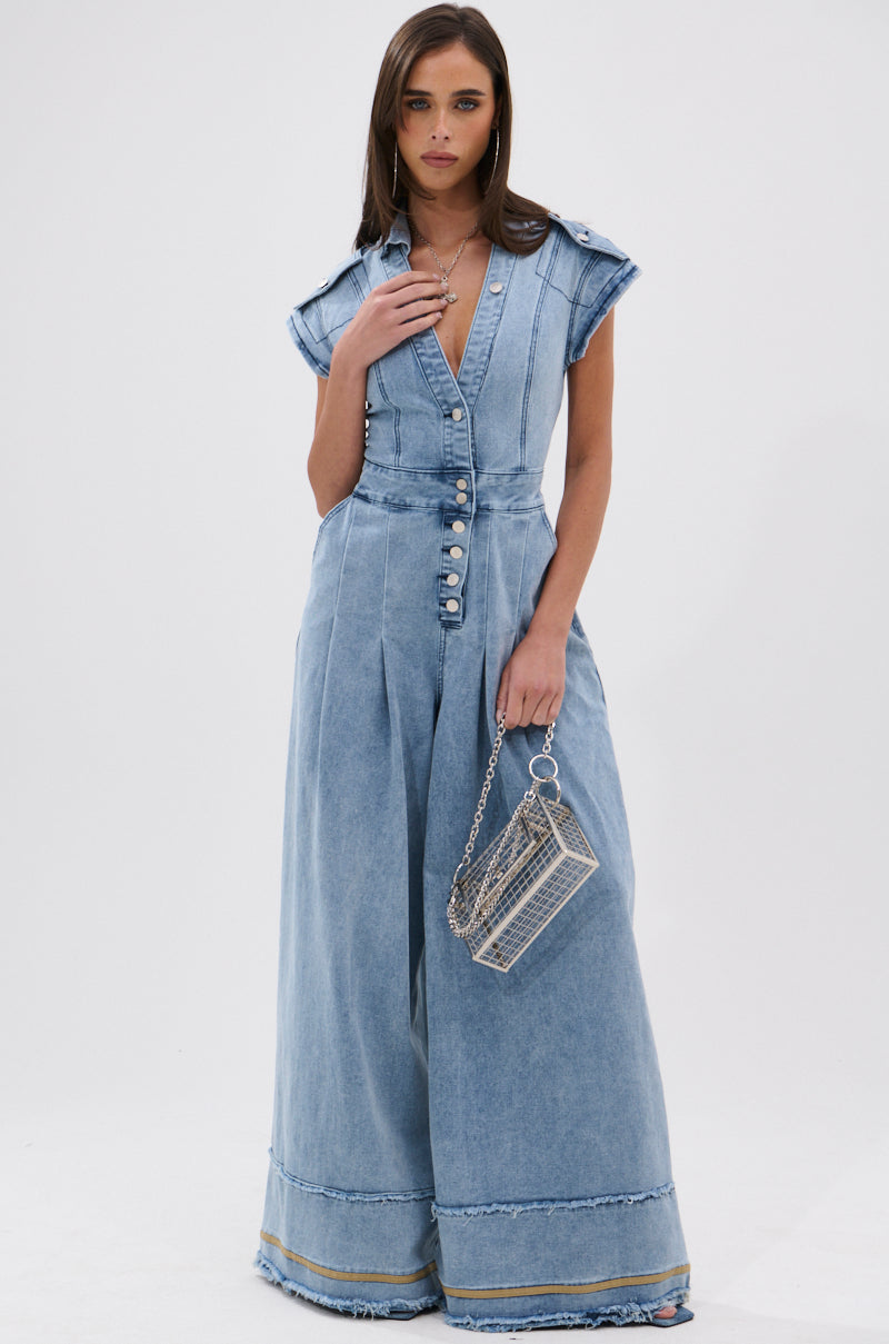 A woman models the AT LEAST I LOOK CUTE DENIM JUMPSUIT, featuring light-wash denim, cap sleeves, a button front, and frayed hems. She holds a silver chain handbag and stands against a plain white background with straight brown hair.