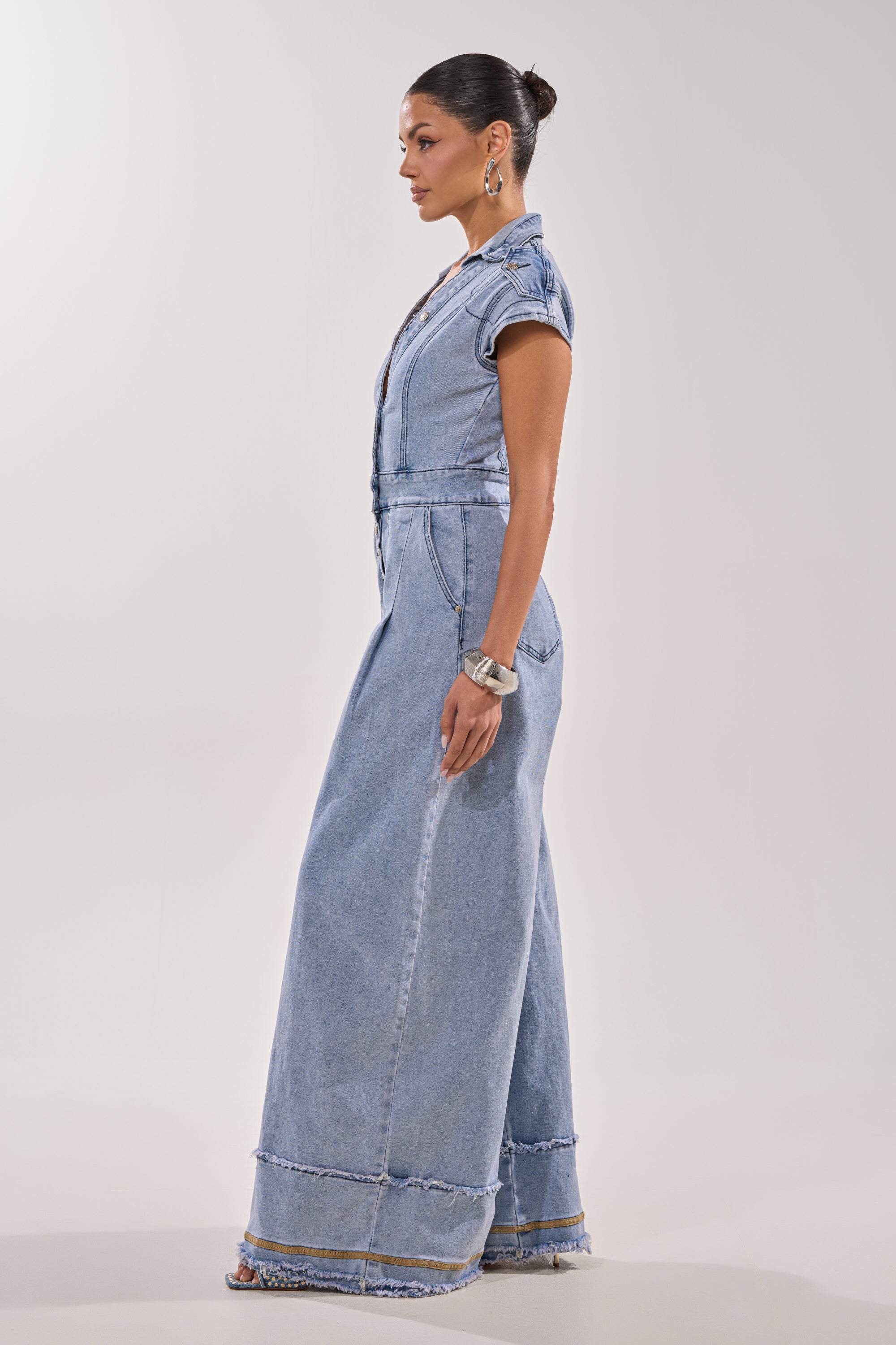 A woman with dark hair in a bun models the AT LEAST I LOOK CUTE DENIM JUMPSUIT, featuring a light wash, wide legs, short sleeves, and frayed hems. She accessorizes with hoop earrings and a bracelet against a plain light background. 4