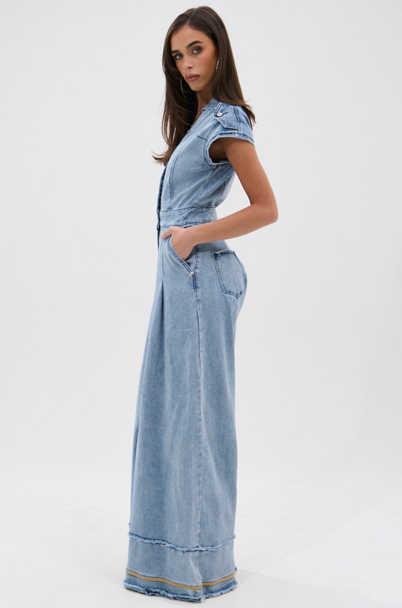 A woman stands sideways with hands in her pockets, wearing the AT LEAST I LOOK CUTE DENIM JUMPSUIT—a light-wash, short-sleeve, full-length collared denim piece with a slightly flared hem—against a plain white background.