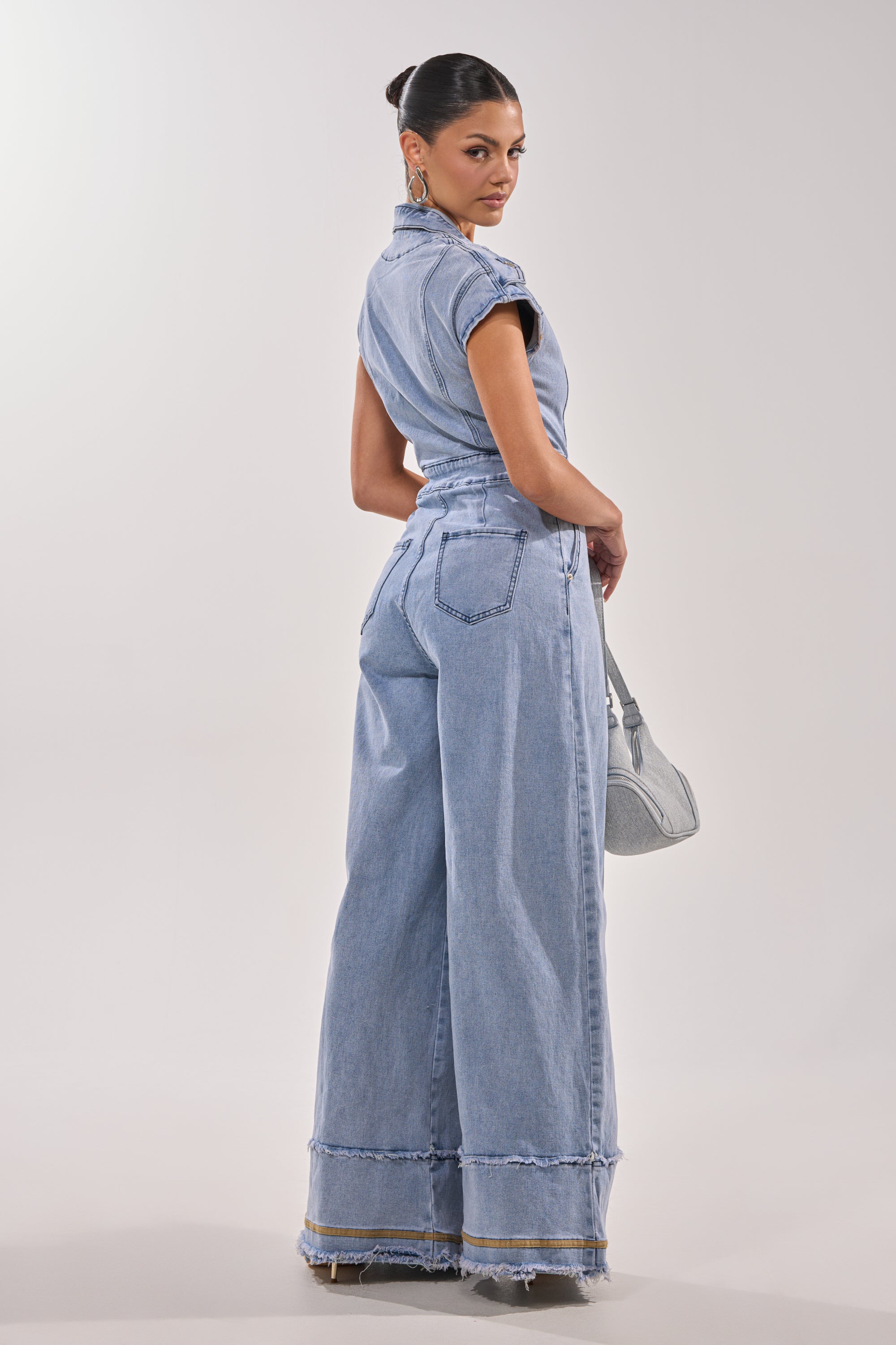 A woman poses in the AT LEAST I LOOK CUTE DENIM JUMPSUIT—light blue with wide legs and frayed hems—holding a gray handbag, her hair in a sleek bun, all set against a plain light background. 5