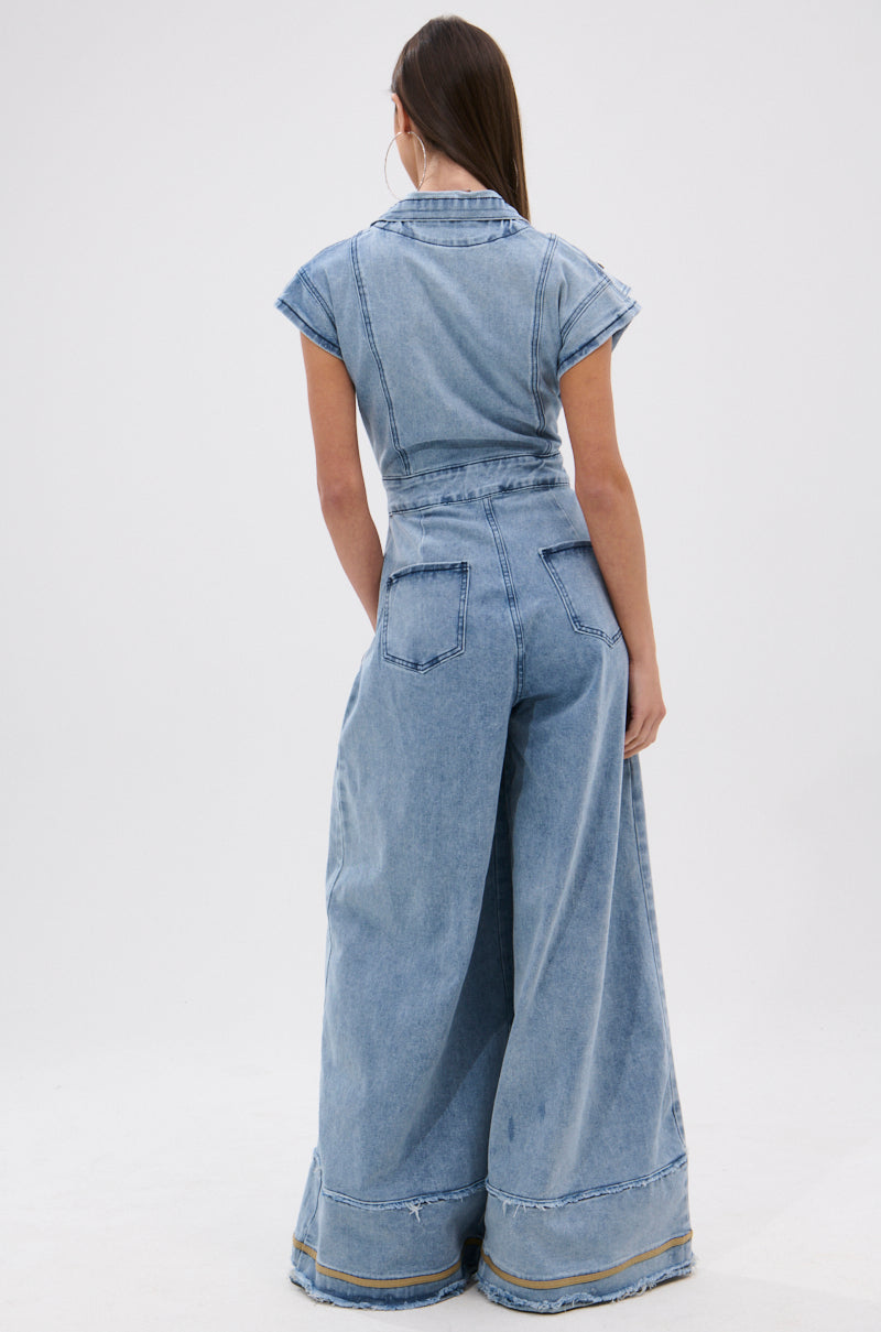 A woman with straight brown hair faces away, modeling the AT LEAST I LOOK CUTE DENIM JUMPSUIT—a utility-chic, light-wash denim piece with wide legs, short sleeves, and two back pockets—set against a plain white background.