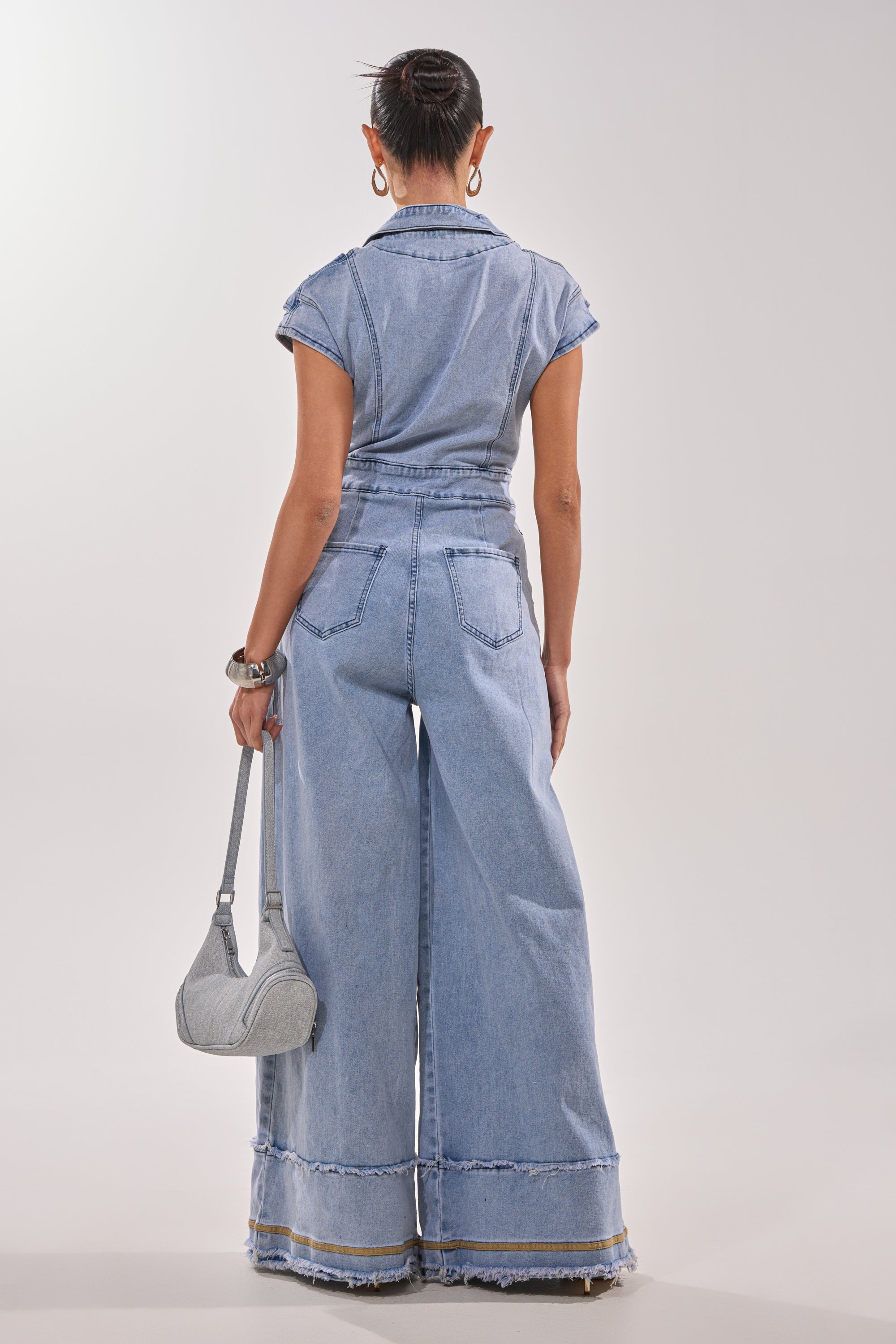 A woman faces away wearing the AT LEAST I LOOK CUTE DENIM JUMPSUIT, featuring light-wash denim and wide-leg frayed hem pants. She carries a silver handbag, with her hair in a bun, accessorized by hoop earrings and a watch. 6