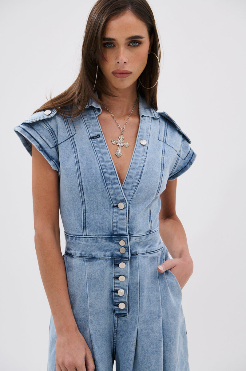 A woman with long brown hair models the AT LEAST I LOOK CUTE DENIM JUMPSUIT, featuring a light wash, short sleeves, silver buttons, and a deep V-neck. She completes the look with hoop earrings and a cross necklace against a white background.