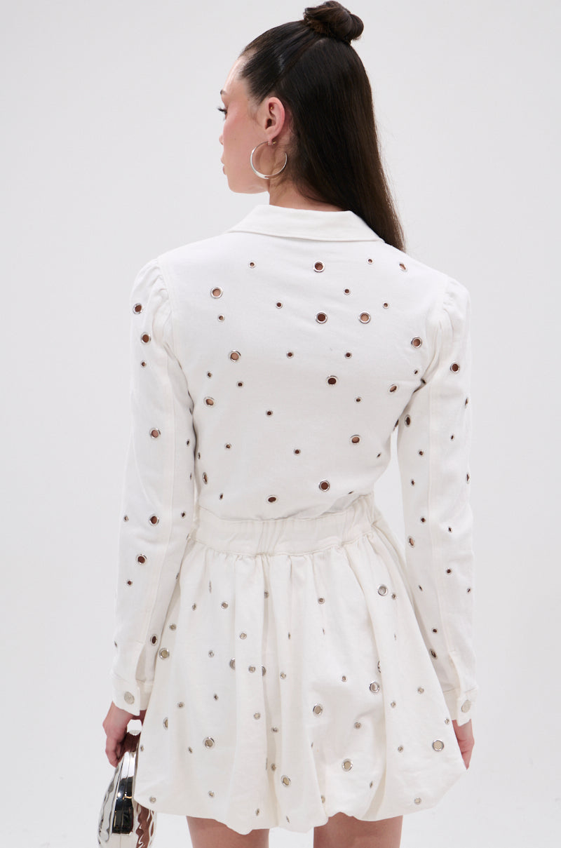 A woman with long dark hair in a half-up bun faces away from the camera, wearing the CUT IT OUT DENIM MINI DRESS WITH GROMMETS—white, long-sleeved, with grommet hardware and a gathered waist—and holds a small silver handbag.
