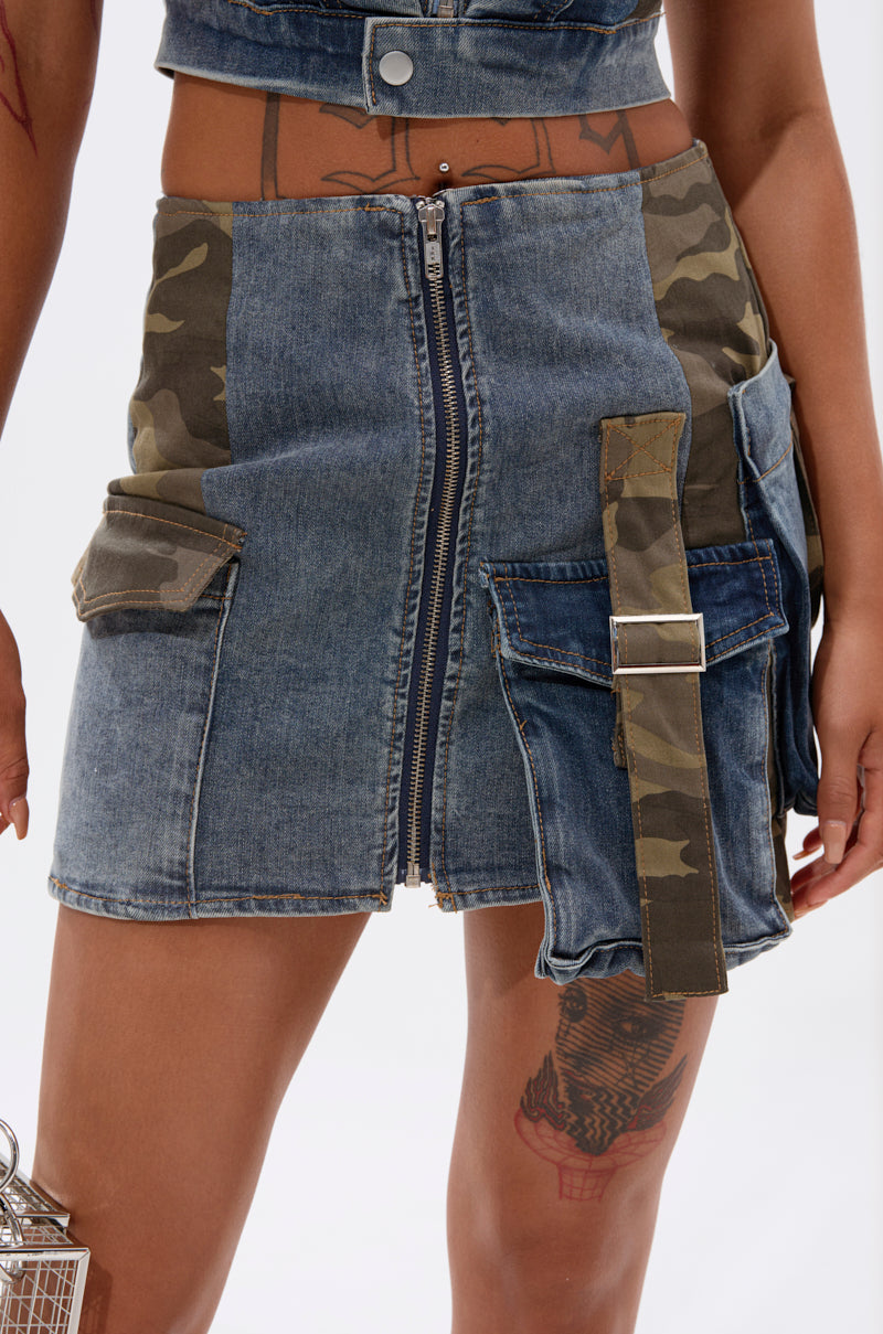 A close-up of the DOWN AND DIRTY CAMO DENIM MINI SKIRT featuring cargo straps, pocket accents, a wide camo buckle strap, and a bold front zipper. The wearer’s tattooed thigh adds an edgy street-style vibe. 0