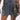 A close-up of the DOWN AND DIRTY CAMO DENIM MINI SKIRT featuring cargo straps, pocket accents, a wide camo buckle strap, and a bold front zipper. The wearer’s tattooed thigh adds an edgy street-style vibe.