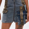 A close-up of the DOWN AND DIRTY CAMO DENIM MINI SKIRT featuring cargo straps, pocket accents, a wide camo buckle strap, and a bold front zipper. The wearer’s tattooed thigh adds an edgy street-style vibe.