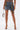 Wearing the DOWN AND DIRTY CAMO DENIM MINI SKIRT with a front zipper and belt details, they pair it with a boxy handbag, clear high heels, and display a skull tattoo on one thigh for an edgy cargo-inspired look.