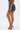 A woman wearing the DOWN AND DIRTY CAMO DENIM MINI SKIRT, clear high-heeled mules, and a woven bag stands against a plain white background.