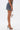 A person in profile wears the DOWN AND DIRTY CAMO DENIM MINI SKIRT, paired with high-heeled clear mules and a small transparent handbag, all set against a plain white background.