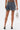 Seen from the back, a woman wears the DOWN AND DIRTY CAMO DENIM MINI SKIRT with a camouflage pocket, high heels, and a silver chain handbag. Her visible tattoos highlight her bold street style.