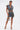 A person stands confidently in the DOWN AND DIRTY CAMO DENIM MINI SKIRT, featuring large pockets and a front zipper. They pair it with a clear purse and clear heels against a plain white background.