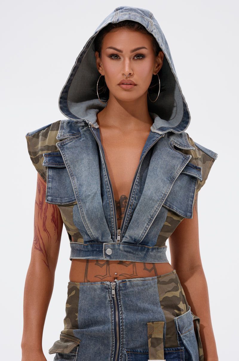 A woman wears the DOWN AND DIRTY DENIM HOODED VEST with matching camo high-waisted pants and hoop earrings, standing confidently against a plain light background. Tattoos decorate her arm. 0