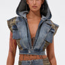 A woman wears the DOWN AND DIRTY DENIM HOODED VEST with matching camo high-waisted pants and hoop earrings, standing confidently against a plain light background. Tattoos decorate her arm.