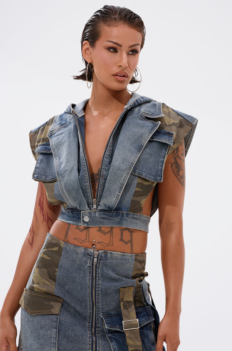 A person with wet-look short hair wears the DOWN AND DIRTY DENIM HOODED VEST in a camo print, styled as a crop top with large pockets, paired with a matching high-waisted skirt. Tattoos and a belly piercing add an edgy vibe. 3