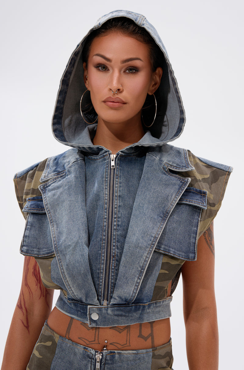 A woman with hoop earrings and facial piercings poses confidently against a plain, light background wearing the DOWN AND DIRTY DENIM HOODED VEST featuring camo details. 4