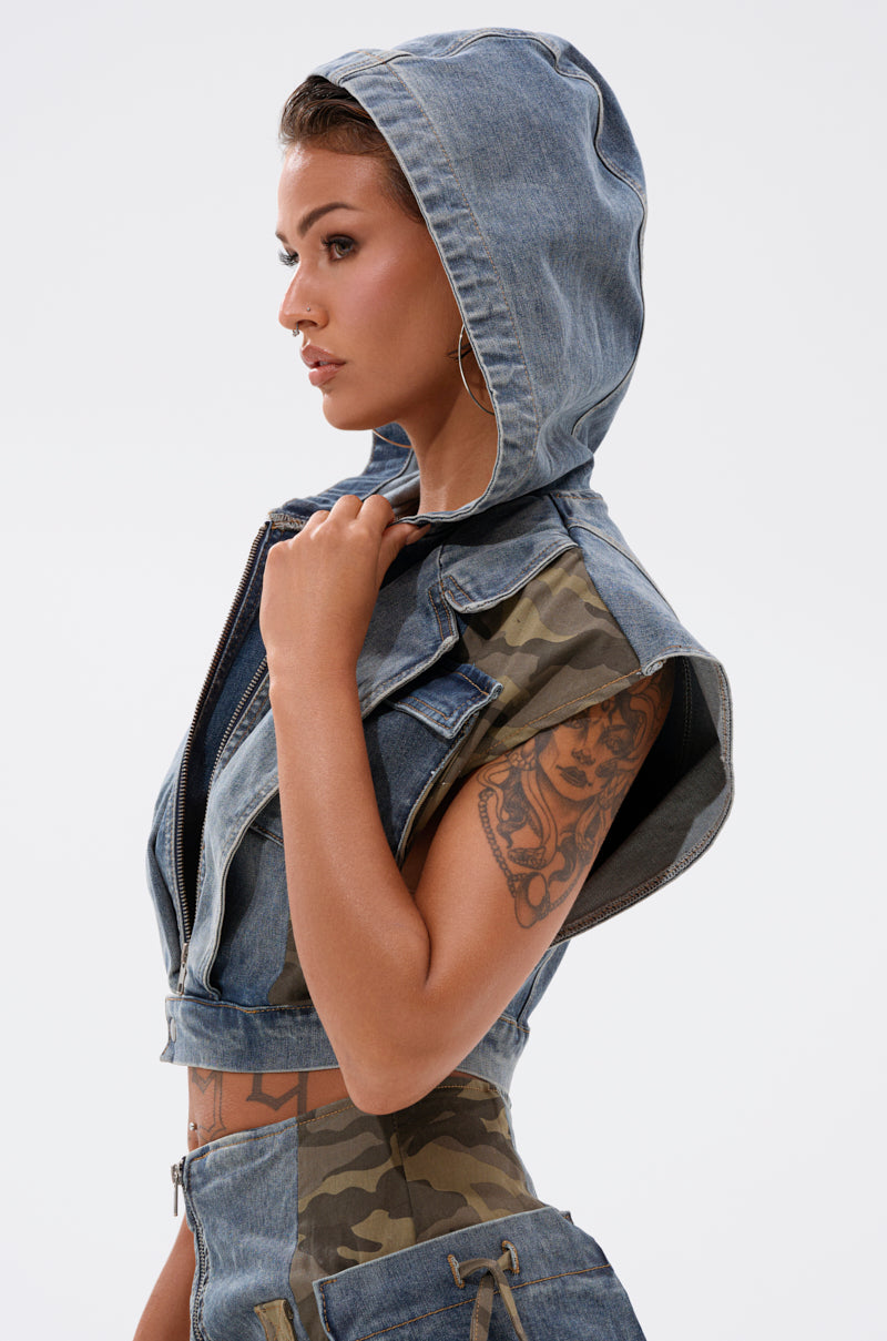 A woman in the DOWN AND DIRTY DENIM HOODED VEST and a camouflage crop top poses in profile against a white background, displaying her upper arm tattoo and wearing hoop earrings. 5