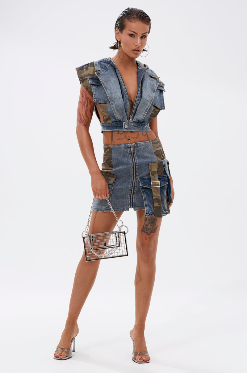 A woman poses confidently in the DOWN AND DIRTY DENIM HOODED VEST—a cropped denim vest with camouflage details—paired with a matching mini skirt, clear handbag, and transparent heels against a plain white background. 7