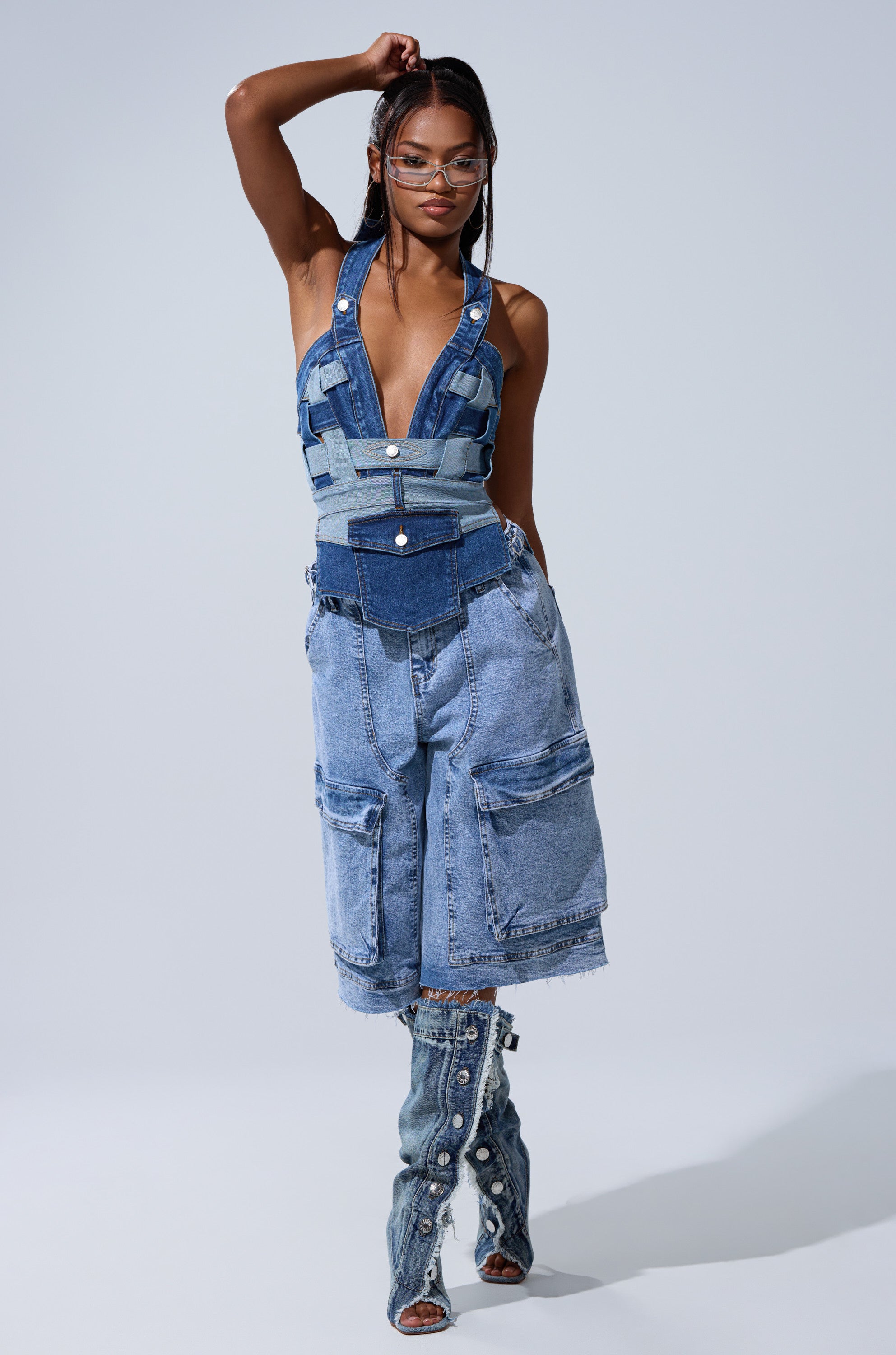 A woman poses in the CATCH ME ABSTRACT DENIM CORSET, styled with oversized denim cargo shorts and pearl-embellished boots. She wears glasses, lifting one arm against a plain backdrop. 4