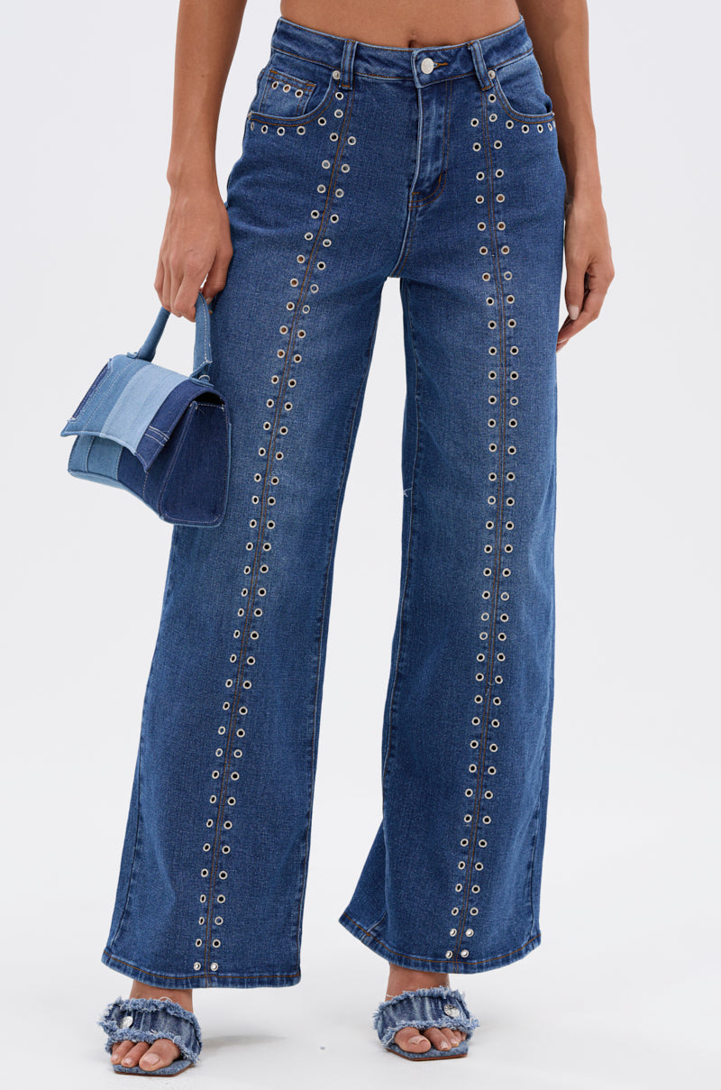 Wearing SOFIA RELAXED DENIM WITH GROMMET—high-rise, wide-leg jeans with vertical metal grommets—a person pairs them with frayed-edge sandals and carries a small denim handbag against a plain white background.
