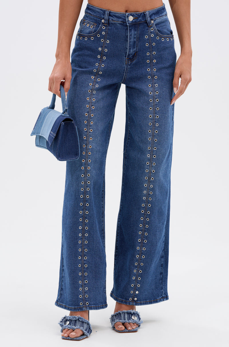 Wearing the SOFIA RELAXED DENIM WITH GROMMET—high-rise blue jeans featuring vertical silver grommets—a person pairs them with a small blue handbag and frayed denim open-toe heels. The wide-leg style pops against the white background.