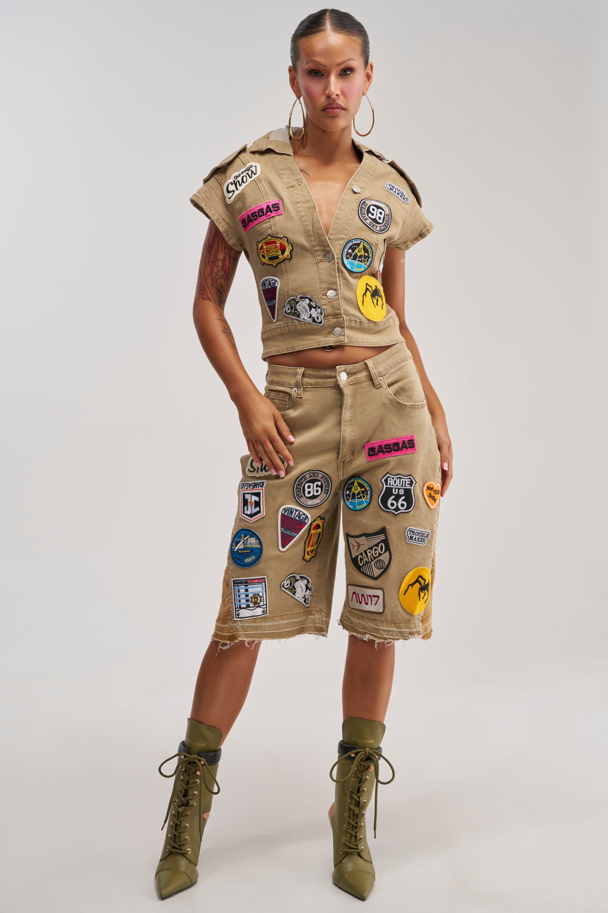 A woman stands confidently wearing a tan, patch-covered vest and the DOWN FOR SOME FUN BERMUDA SHORT—beige denim bermuda shorts with frayed hems and playful, colorful embroidered patches—paired with green lace-up boots. The outfit highlights utility-chic style against a plain background.