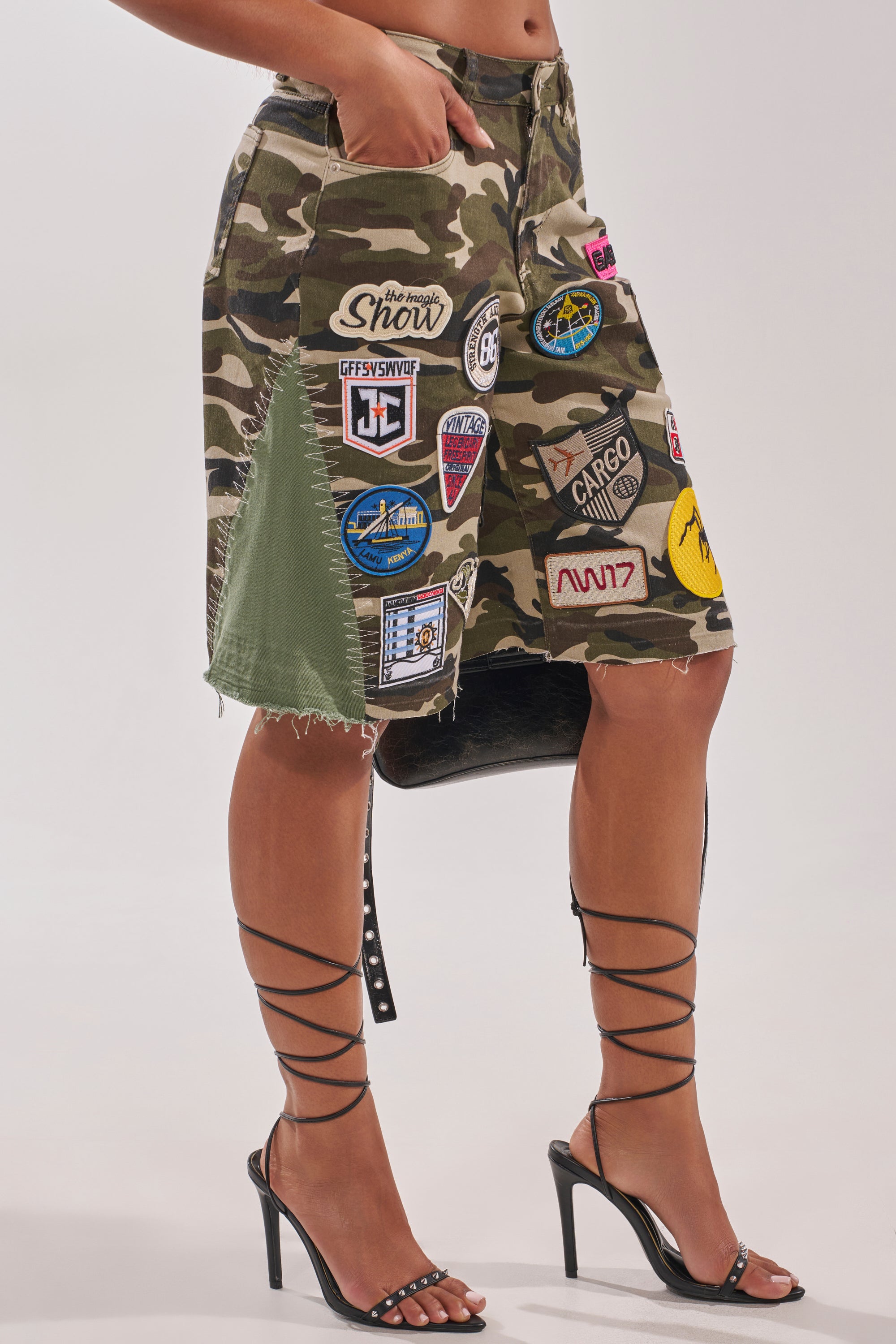Someone wears the DOWN FOR SOME FUN CAMO BERMUDA SHORT—knee-length camo shorts with colorful patches—paired with black strappy high heel sandals, hand in pocket, standing against a plain, light background. 4