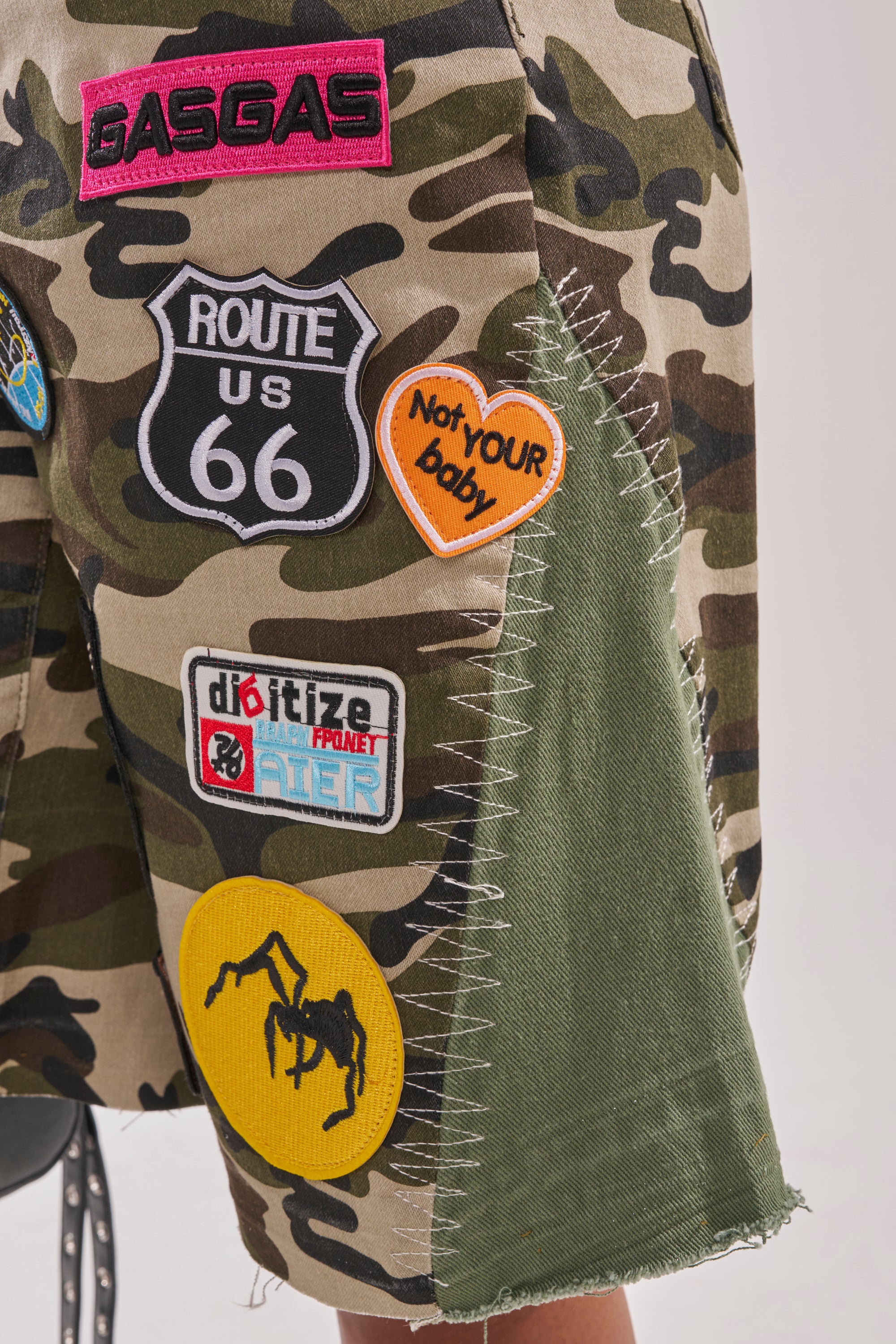 The DOWN FOR SOME FUN CAMO BERMUDA SHORT showcases bold patchwork details—“GASGAS,” “Route US 66,” an orange “Not YOUR baby” heart, and a yellow spider—pairing standout stitching with streetwear attitude. 7