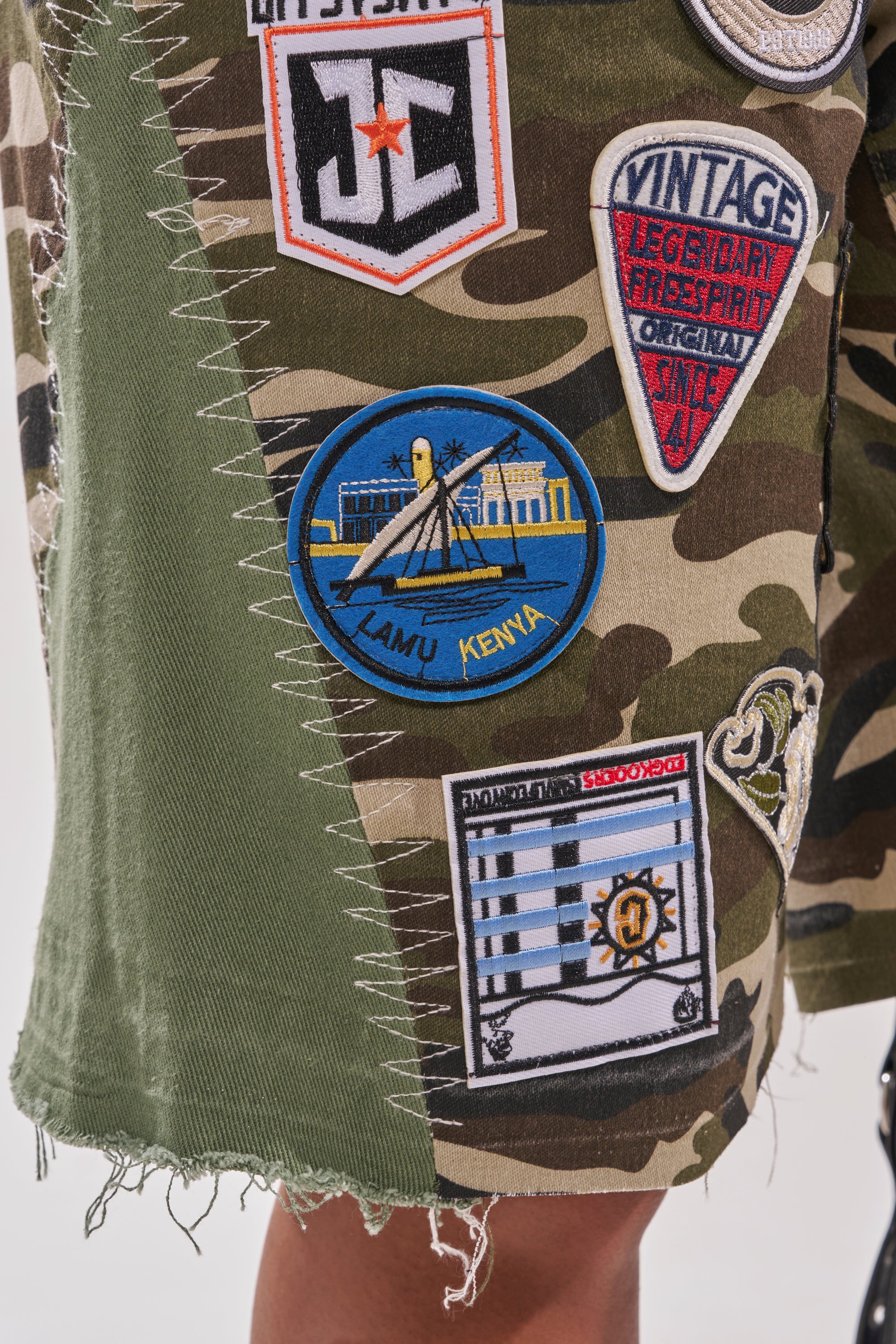 Close-up of the DOWN FOR SOME FUN CAMO BERMUDA SHORT, featuring camo fabric, colorful embroidered patches like a blue "Lamu Kenya" patch and vintage badge. White stitching connects this streetwear piece's panels for a standout look. 8