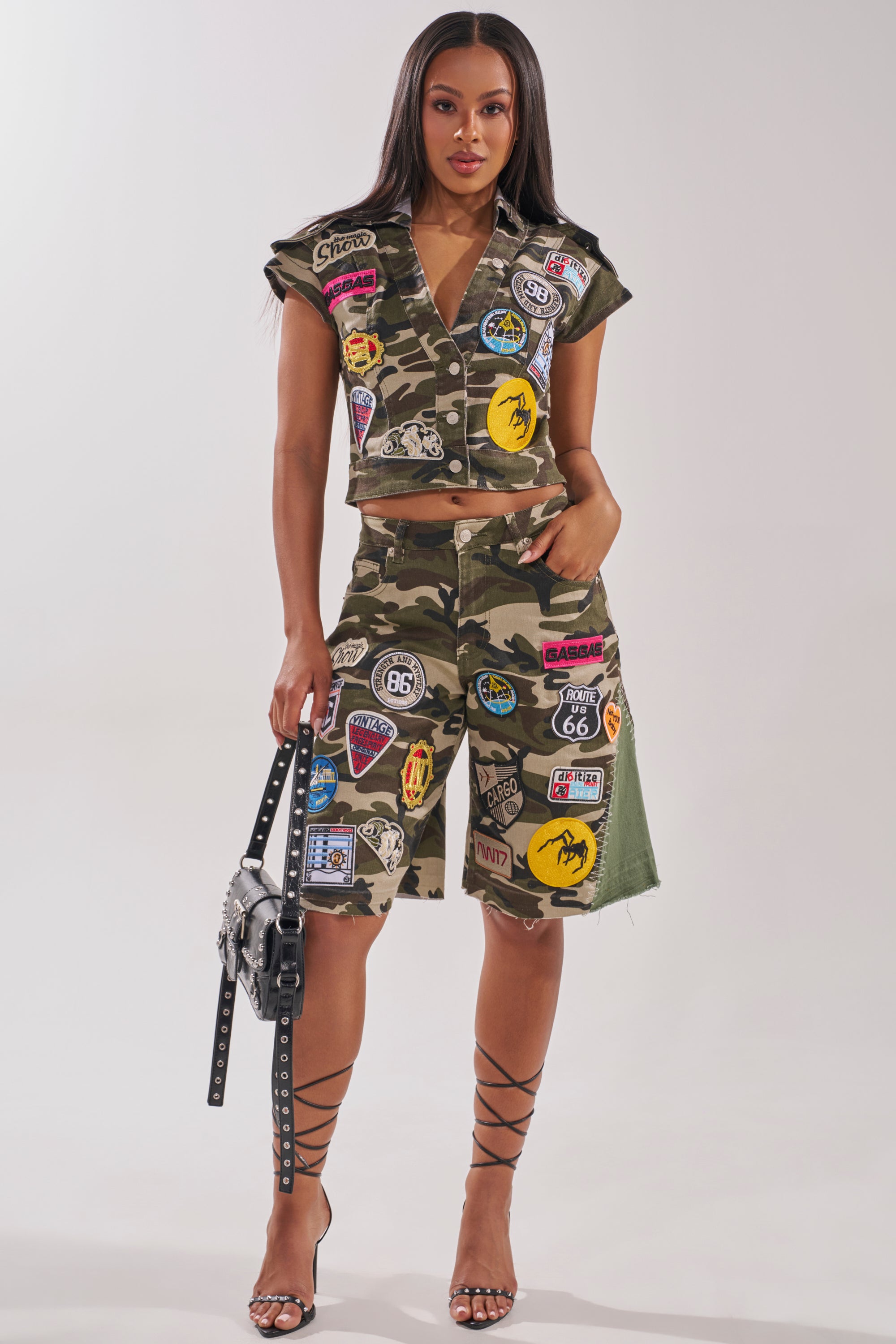 A woman poses in the DOWN FOR SOME FUN CAMO BUTTON DOWN, styled with a camo skirt, colorful patches, a studded handbag, and high-heeled lace-up sandals against a simple light backdrop. 5